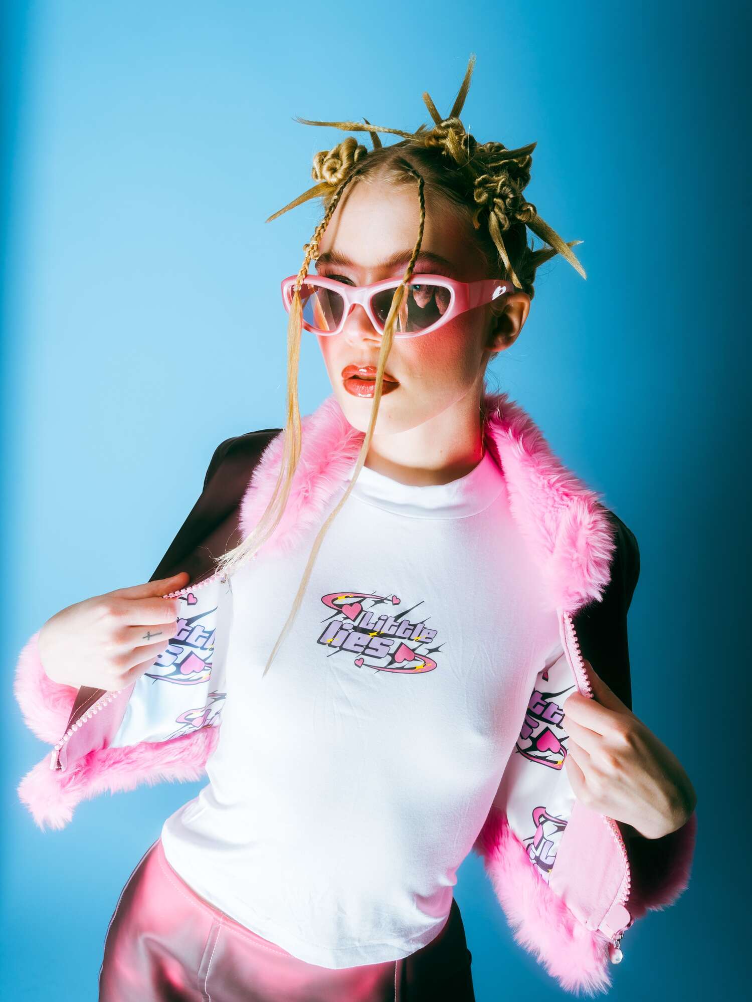 Model wearing a white “Little Lies” graphic tee under a fuchsia faux-fur–trimmed jacket with a sculptural twisted-bun hairstyle and pink cat-eye sunglasses, on bright blue background.