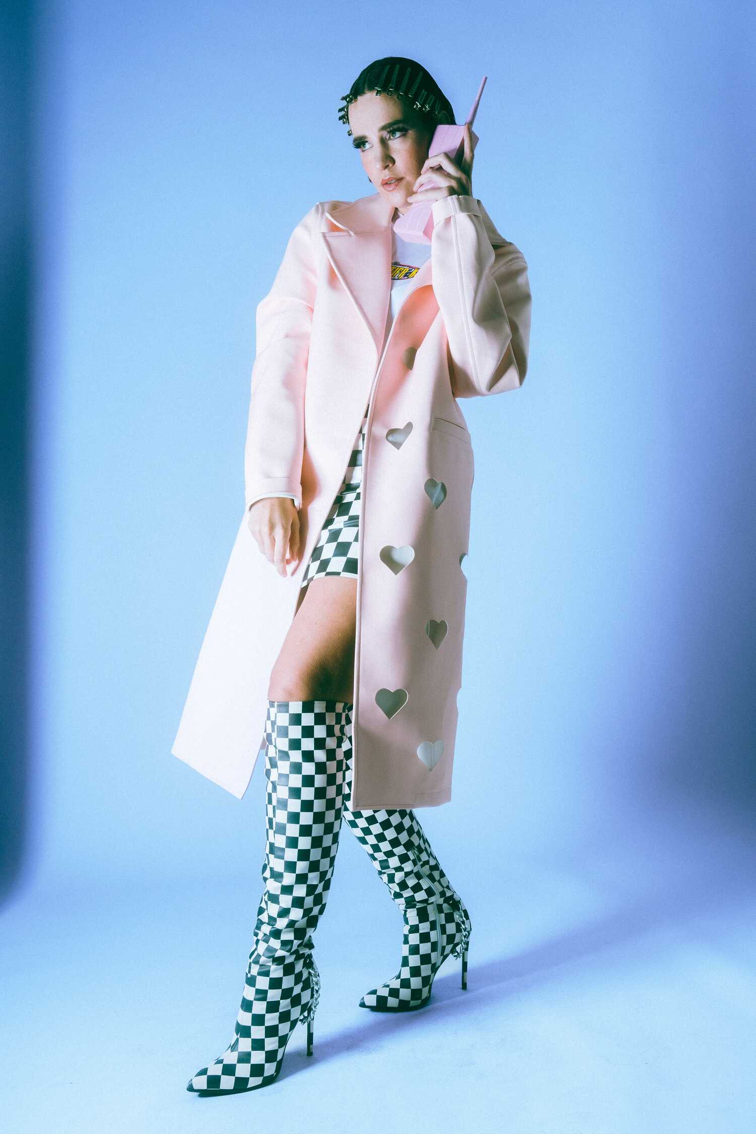 Model in a pastel-pink coat with heart-shaped cutouts, black-and-white checkered knee-high boots, holding a retro pink handset.