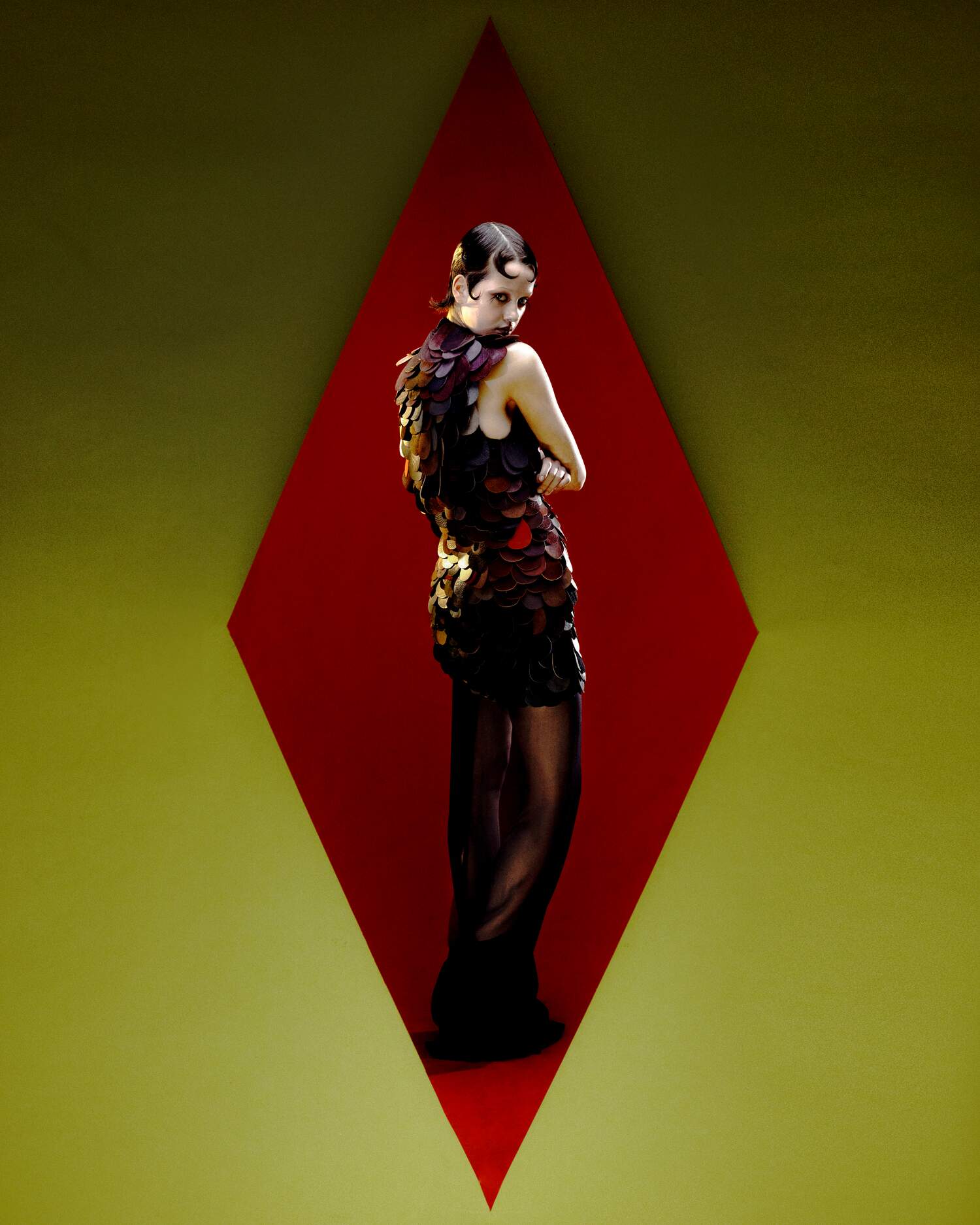 AW25 campaign image: Model wears a sculptural scale-textured mini dress with sheer black hem, framed by a sharp red diamond cutout.
