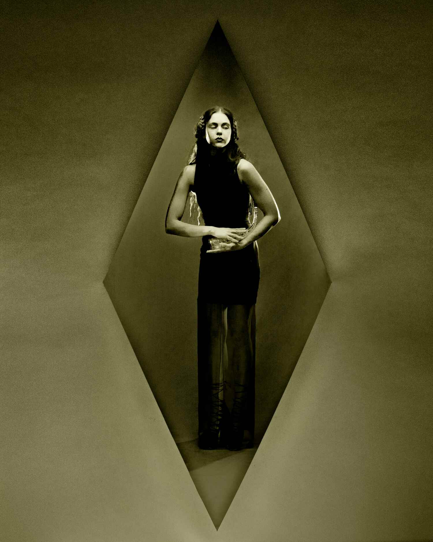 Sepia-toned portrait of model in a sleeveless column dress, framed by a triangular arch and standing still with hands clasped.