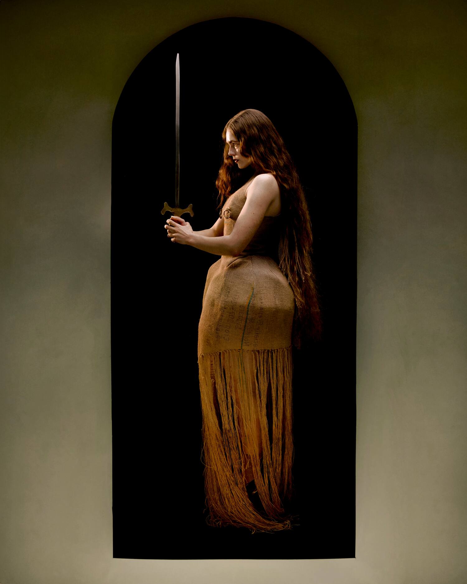 Model holds a long sword while wearing a fringed burlap gown, standing within a dark arch with loose flowing hair.