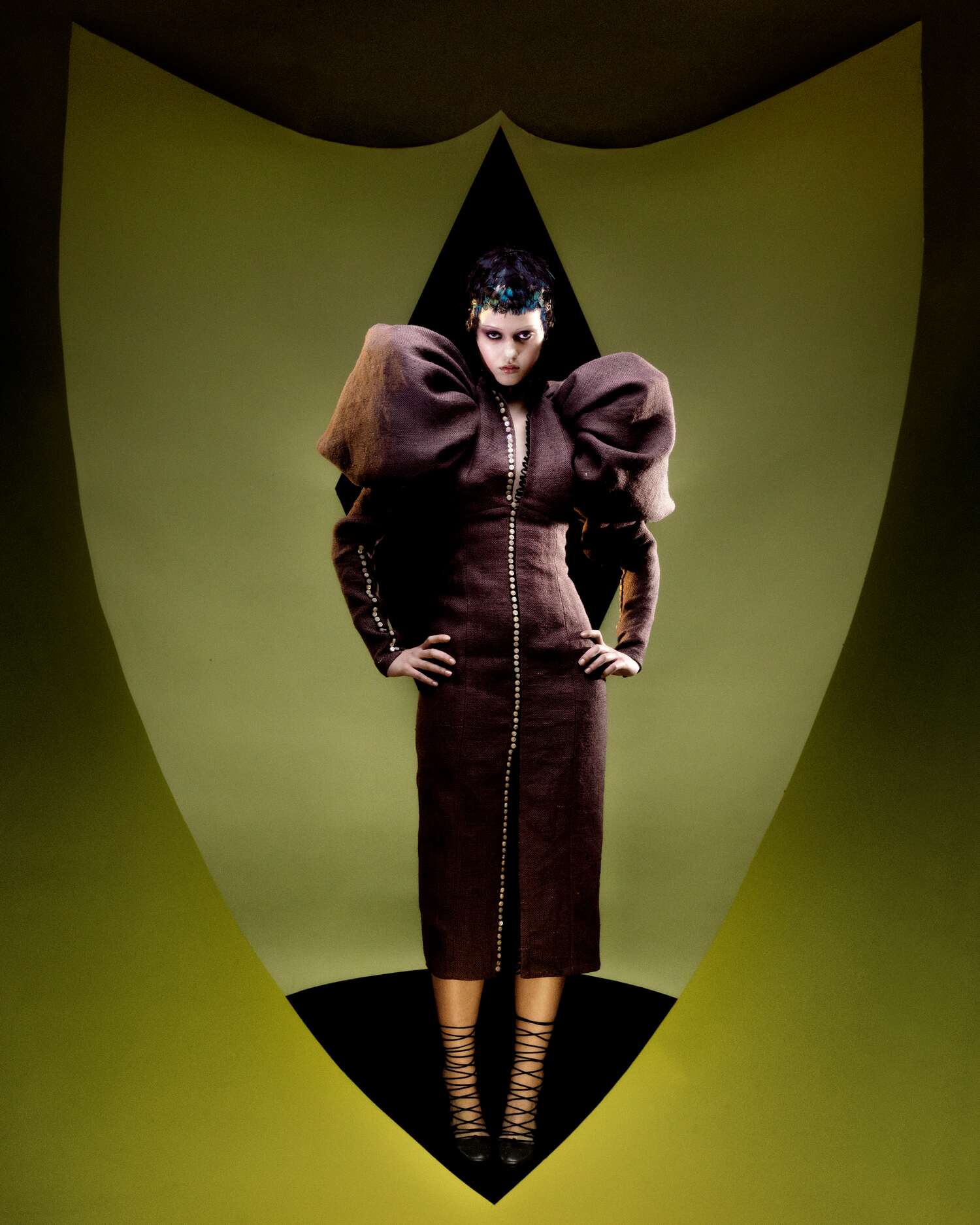 Model stares forward in a structured brown dress with exaggerated shoulders, standing inside a curved green backdrop.