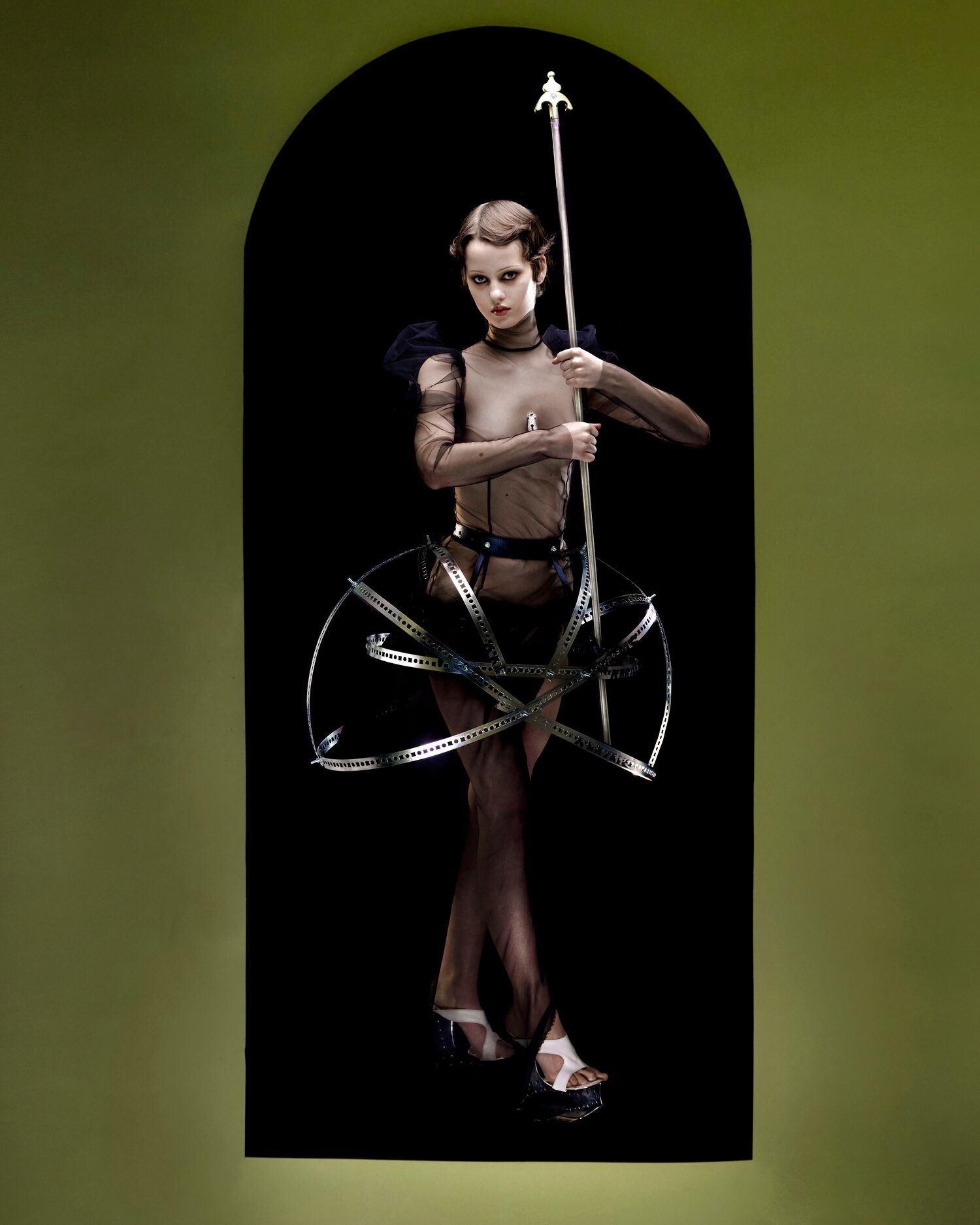 Model in sheer top and metallic cage skirt holds spear — a structured AW25 look staged inside a dark arch.