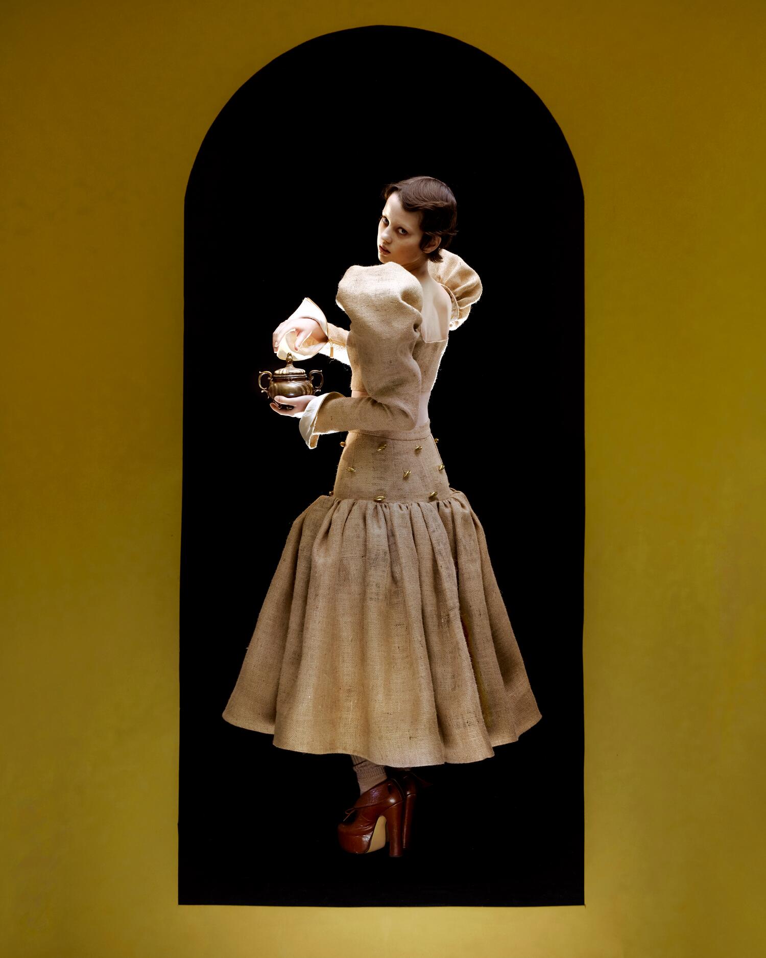 Model holds a brass teapot wearing a burlap-textured beige gown with a voluminous skirt, standing beneath a yellow arch.