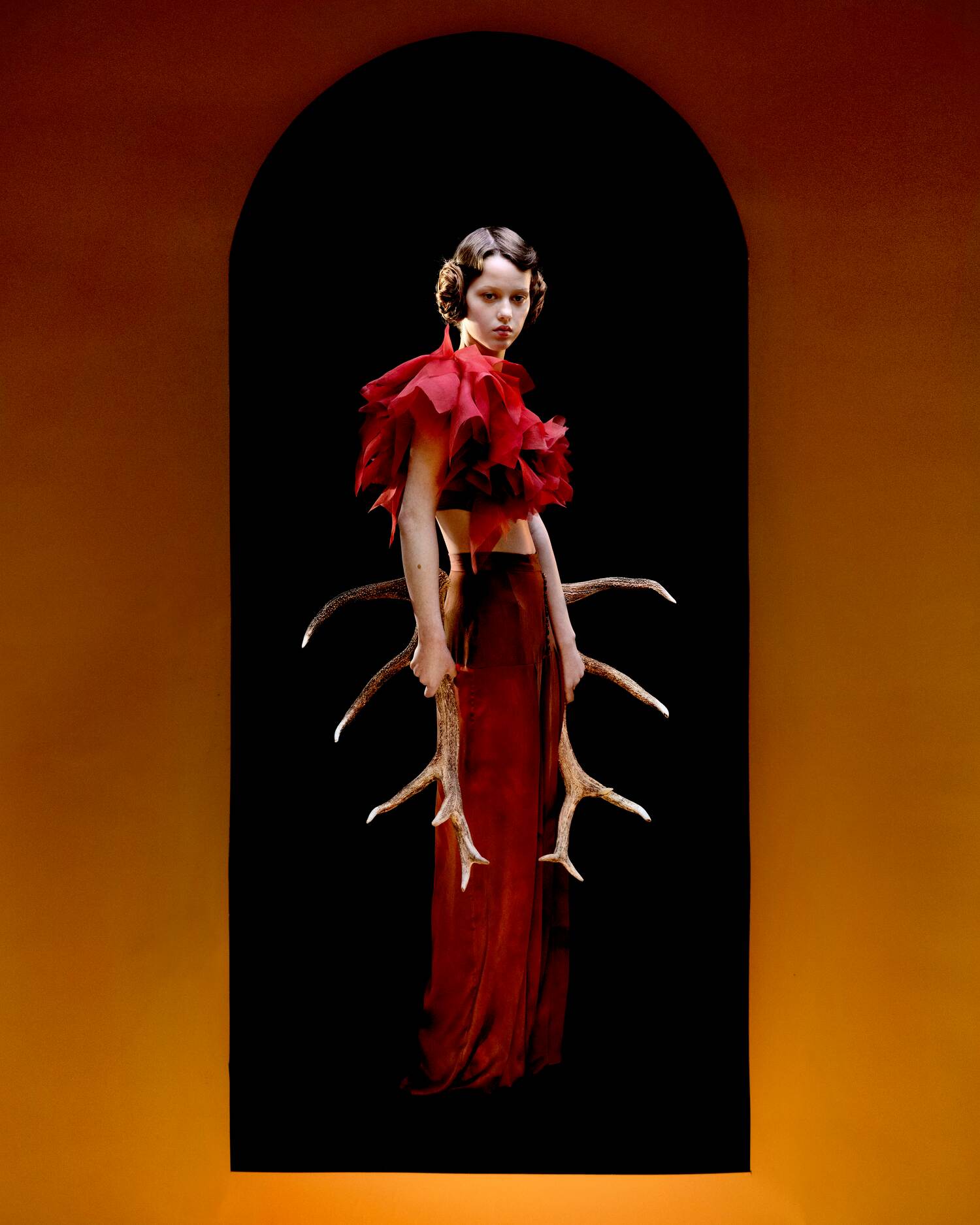 Model in a crimson sculptural top and skirt holds deer antlers, framed by a bold orange backdrop and arched cutout.