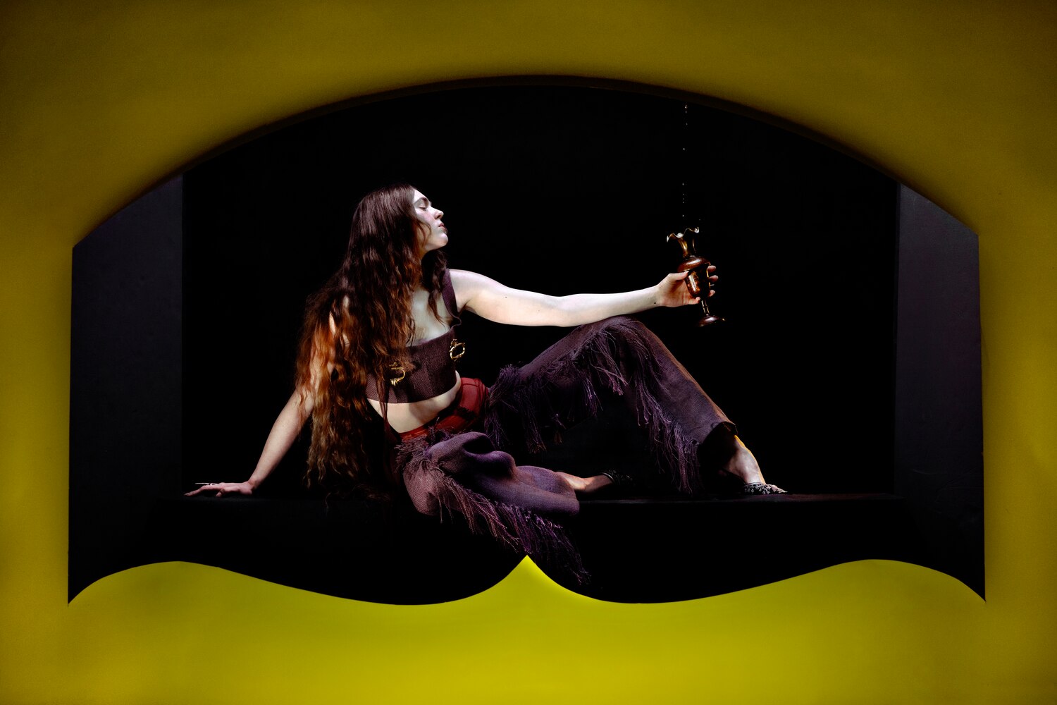 AW25: Model reclines with a brass goblet in hand, wearing fringed dark garments inside a yellow sculptural setting