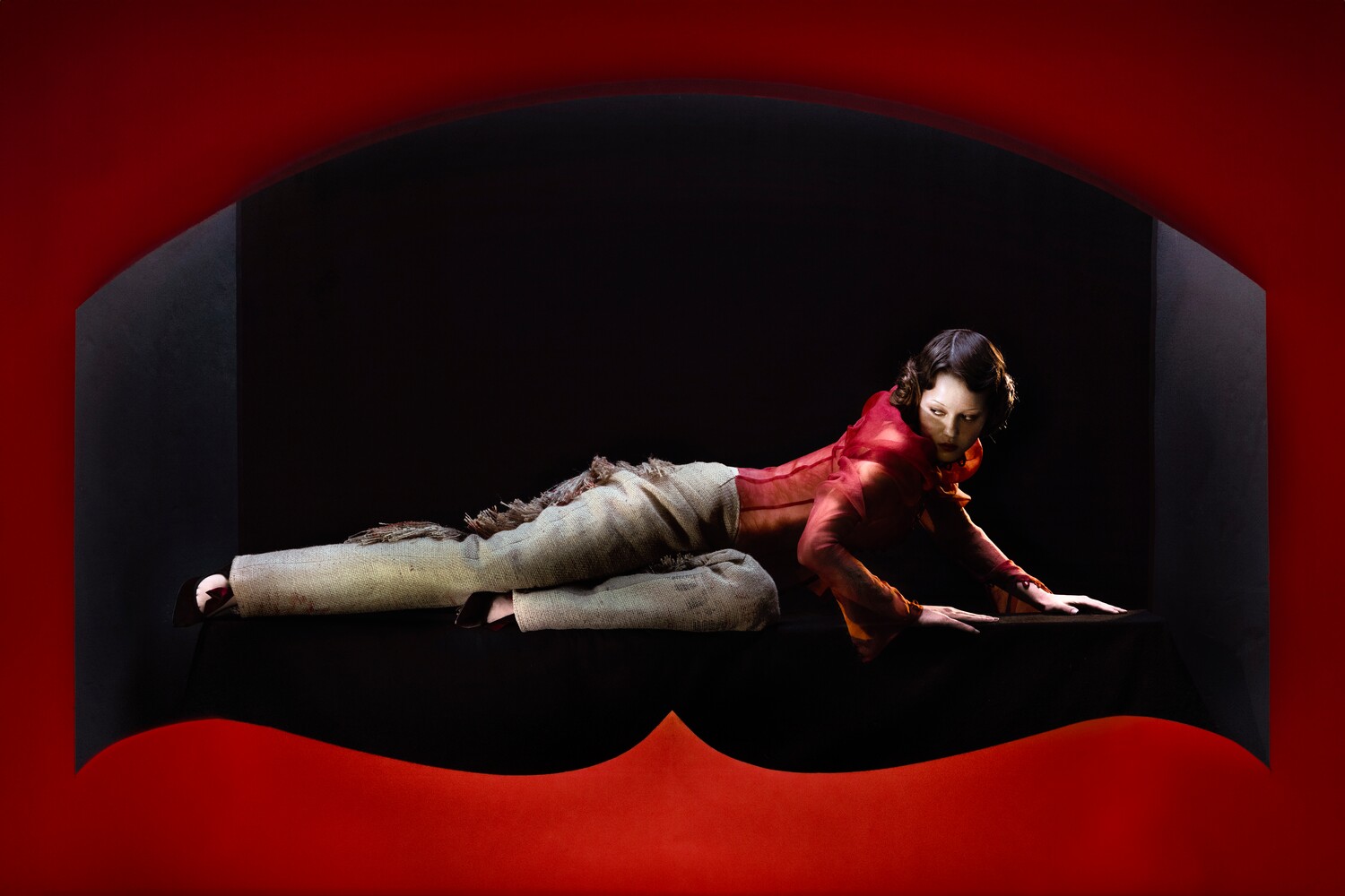 Model in a red corset and burlap-textured trousers reclines across a dark surface, lit by moody red architectural curves.