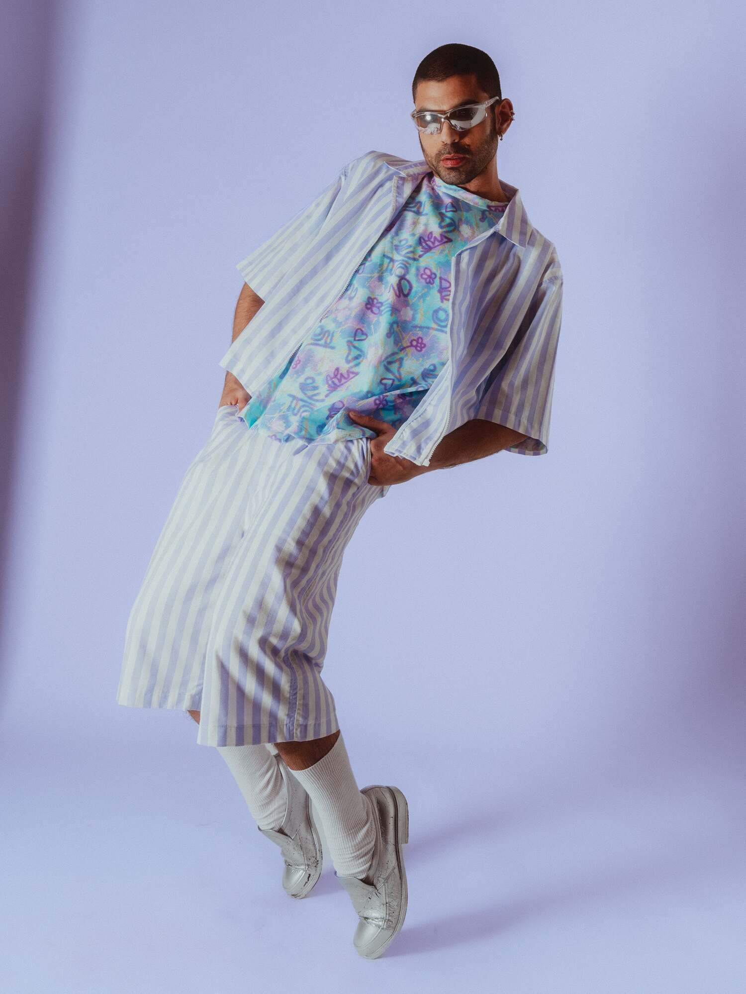 Male model in pastel-striped shirt-and-shorts over a pastel print tee, silver loafers, lavender floor.