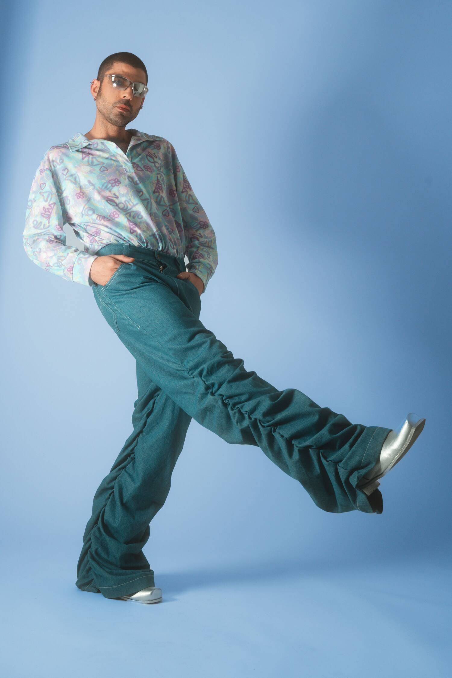 Male model in pastel-print Sunday Morning shirt and scrunched trousers kicking a leg on a light-blue floor.