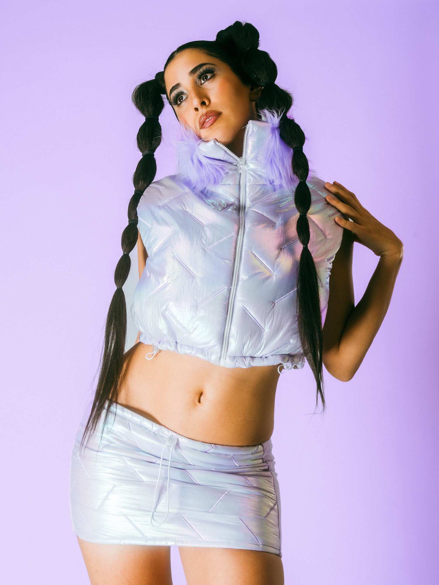 Model in an iridescent lavender quilted vest and matching micro-skirt, hair styled in twin bubble braids, posed against a pastel-purple backdrop.