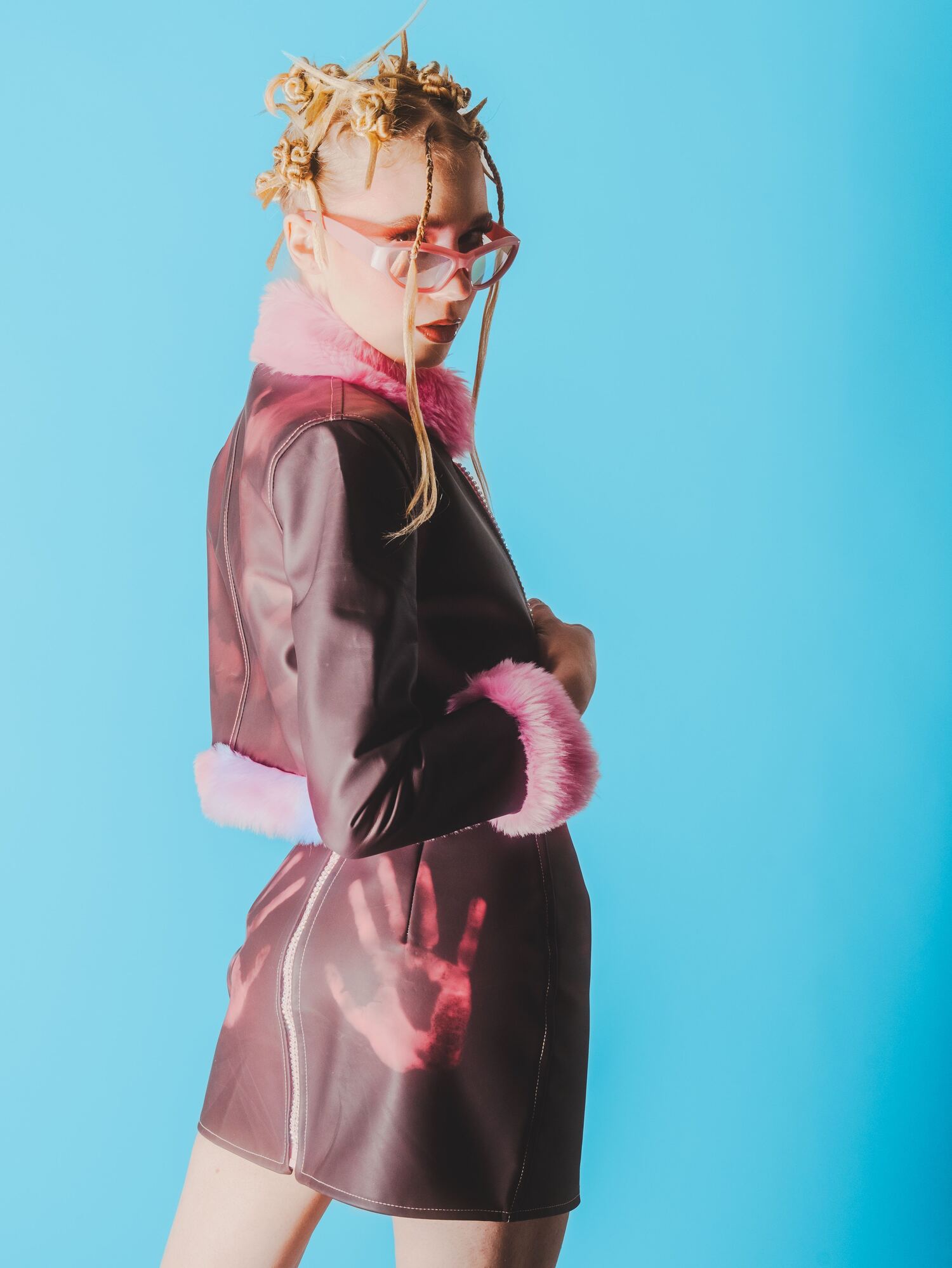 Side profile of model in a deep burgundy faux-leather coat, trimmed with hot-pink faux fur and printed with a subtle handprint detail, set against a sky-blue background.