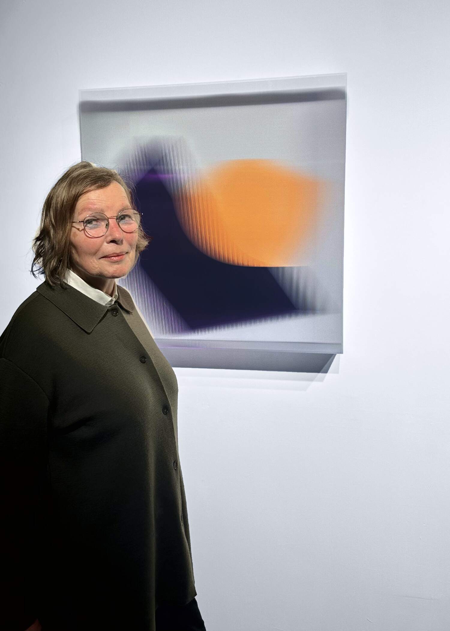 Artist Christiane Grimm standing in front of her glass-based artwork, which features layered reflections and blurred fields of orange, purple, and grey.